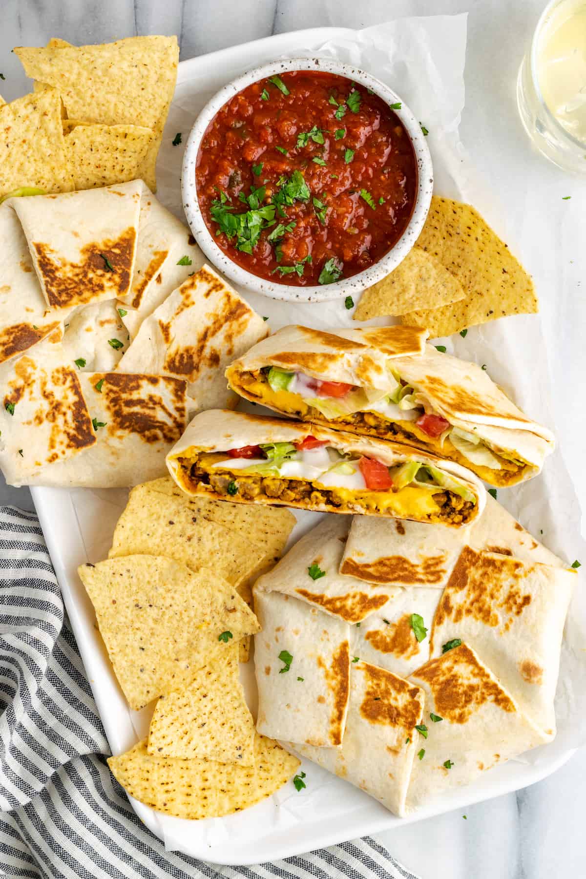 A plate with two whole vegan crunchwrap supremes, one crunchwrap supreme cut in half, some tortilla chips, and a bowl of salsa