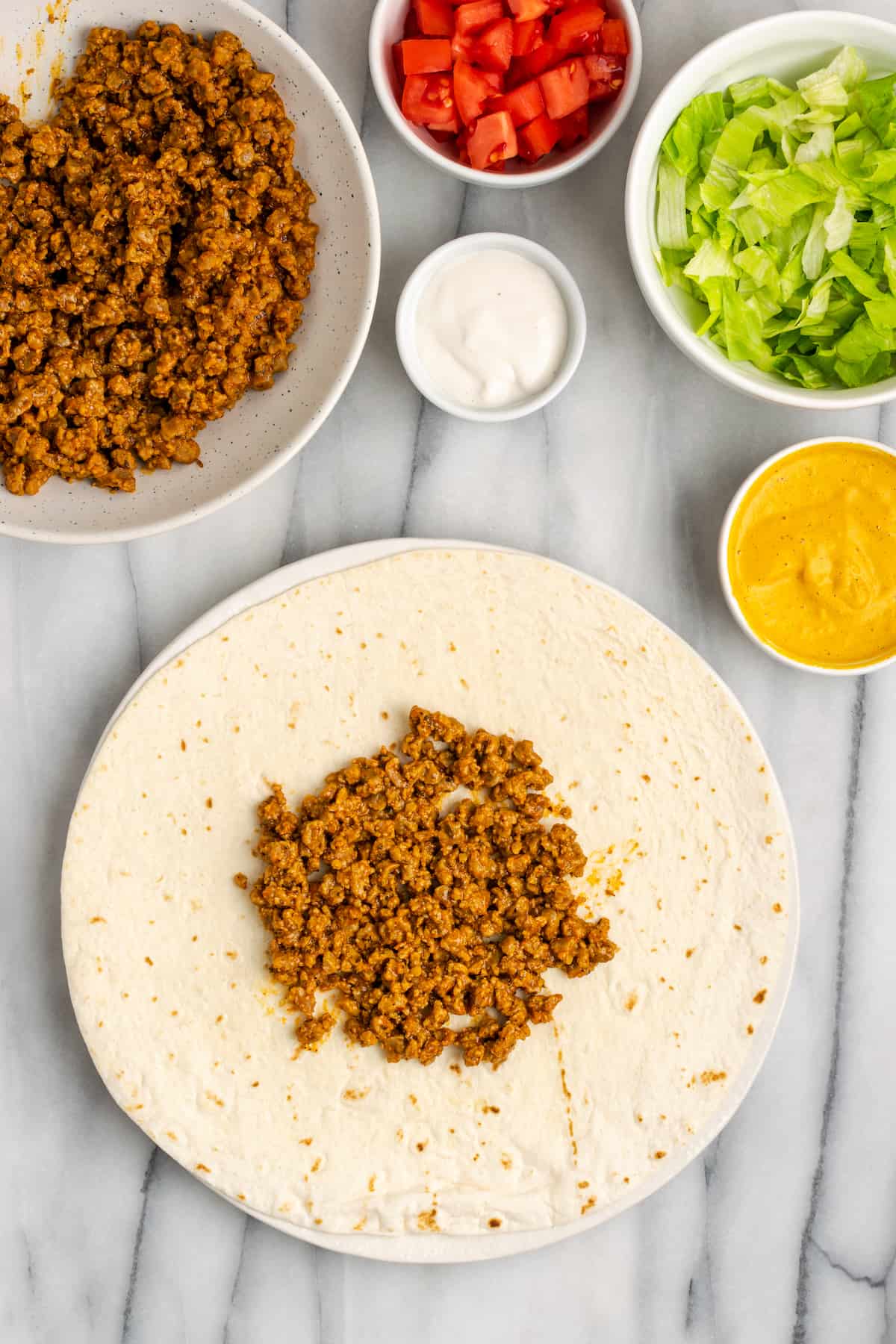A flour tortilla topped with vegan meat crumbles, next to bowls of vegan meat crumbles, vegan cheese sauce, vegan sour cream, lettuce, and tomatoes