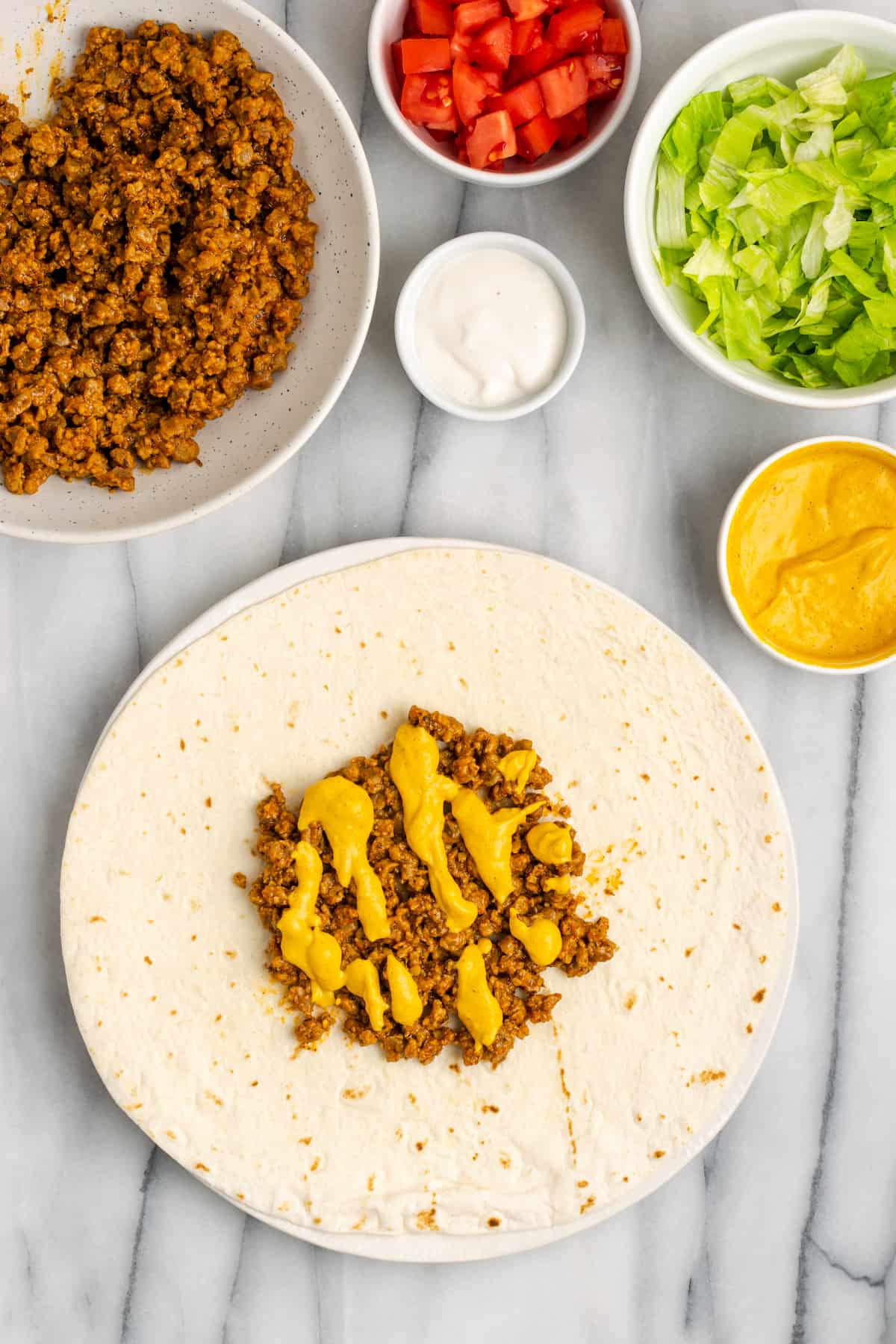 A flour tortilla topped with vegan meat crumbles and vegan cheese sauce, next to bowls of vegan crumbles, vegan cheese sauce, sour cream, lettuce, and tomatoes