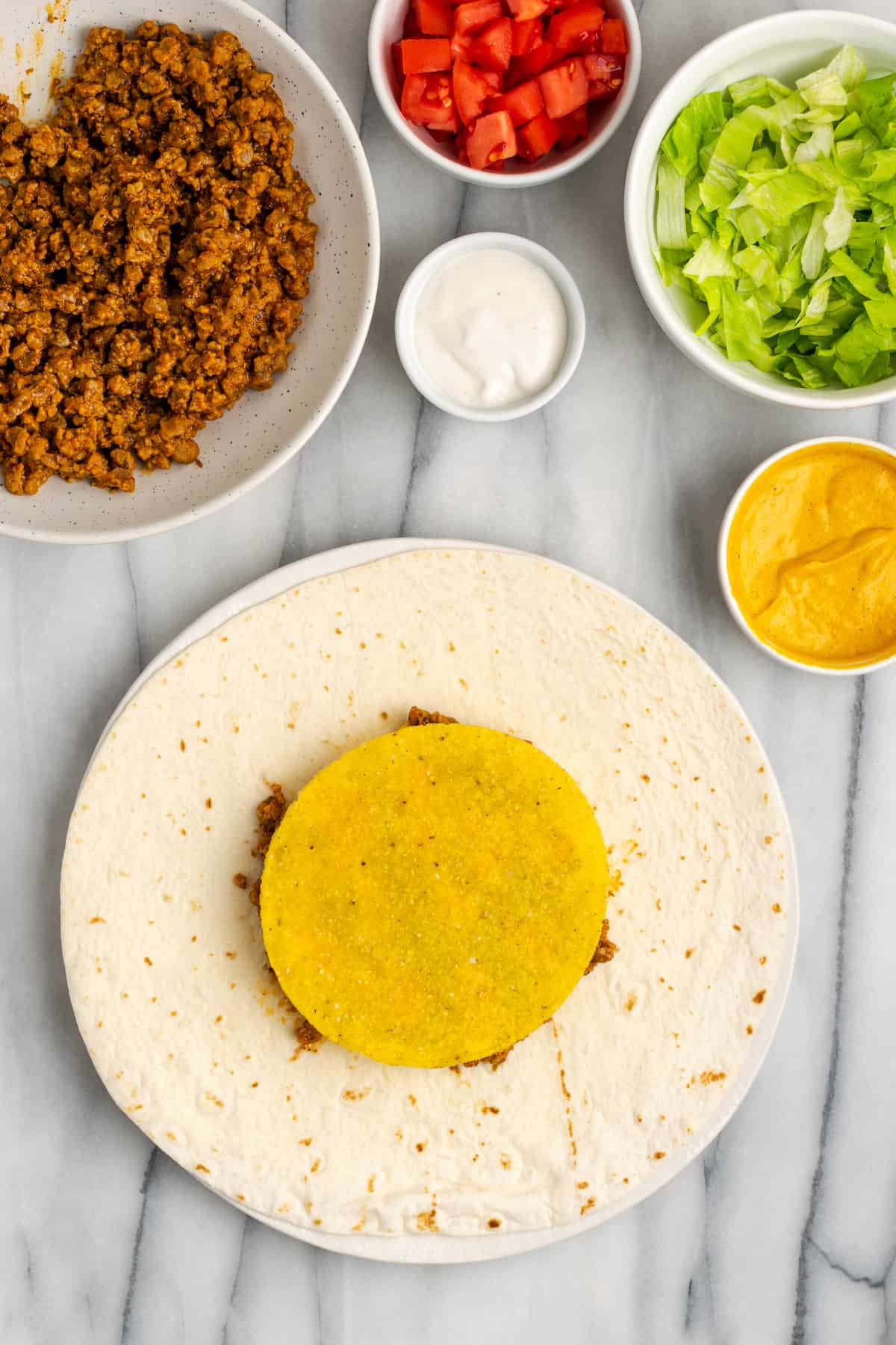 A flour tortilla topped with vegan meat crumbles, vegan cheese sauce, and a tostada, next to bowls of vegan meat crumbles, vegan cheese sauce, vegan sour cream, lettuce, and tomatoes