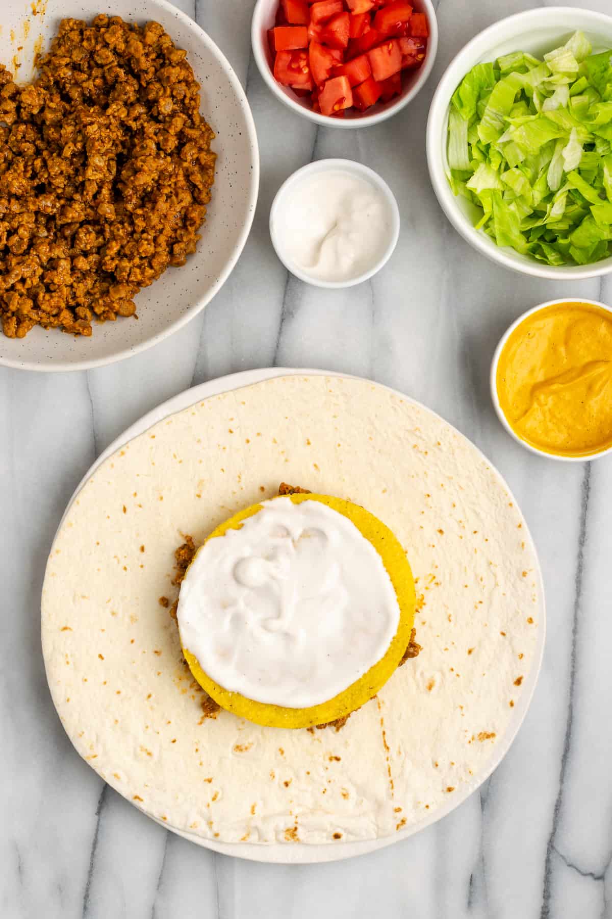 A flour tortilla topped with vegan meat crumbles, vegan cheese sauce, a tostada, and vegan sour cream, next to bowls of meat crumbles, cheese sauce, sour cream, lettuce, and tomatoes