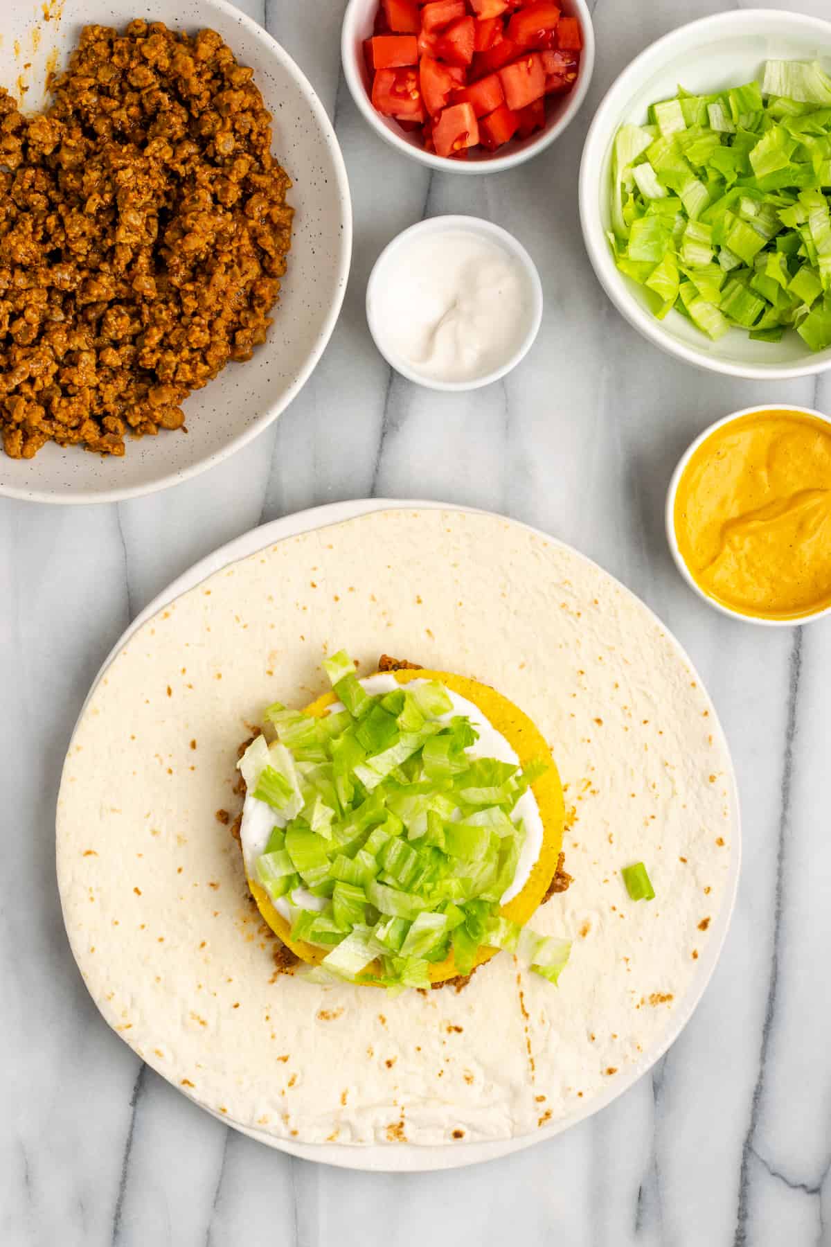 A flour tortilla topped with vegan meat crumbles, vegan cheese sauce, a tostada, vegan sour cream, and lettuce, next to bowls of meat crumbles, sour cream, cheese sauce, lettuce, and tomatoes