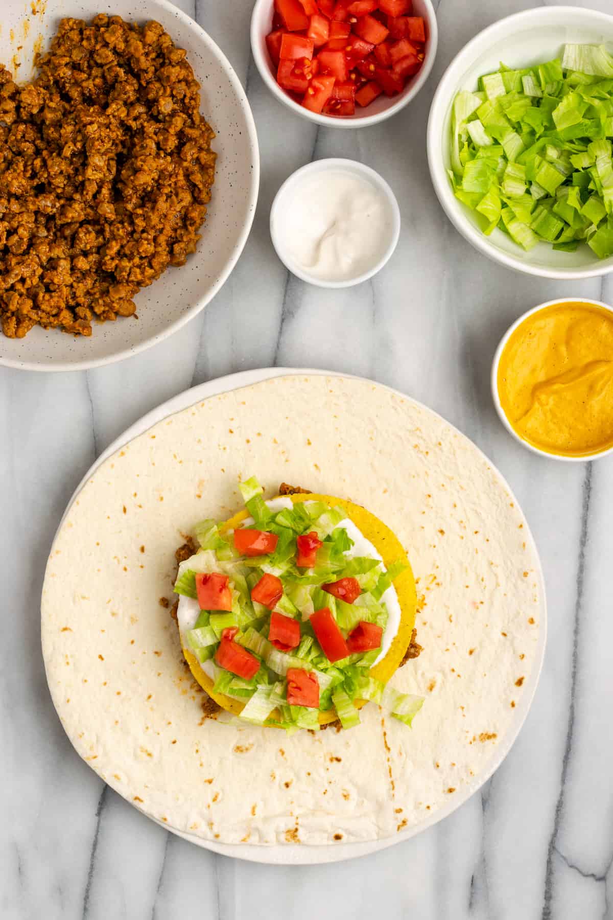 A tortilla topped with vegan meat crumbles, vegan cheese sauce, a tostada, vegan sour cream, lettuce, and tomatoes, next to bowls of meat crumbles, cheese sauce, sour cream, lettuce, and tomatoes