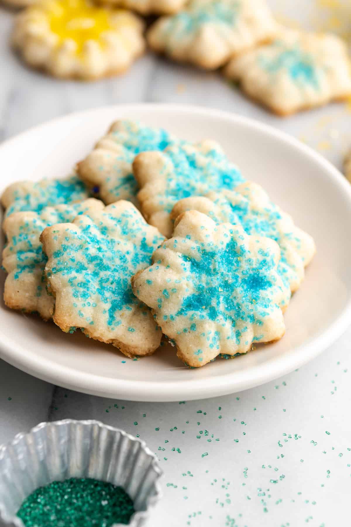 A plate filled with spritz cookies covered in blue sprinkles, with cookies in the background with different sprinkles