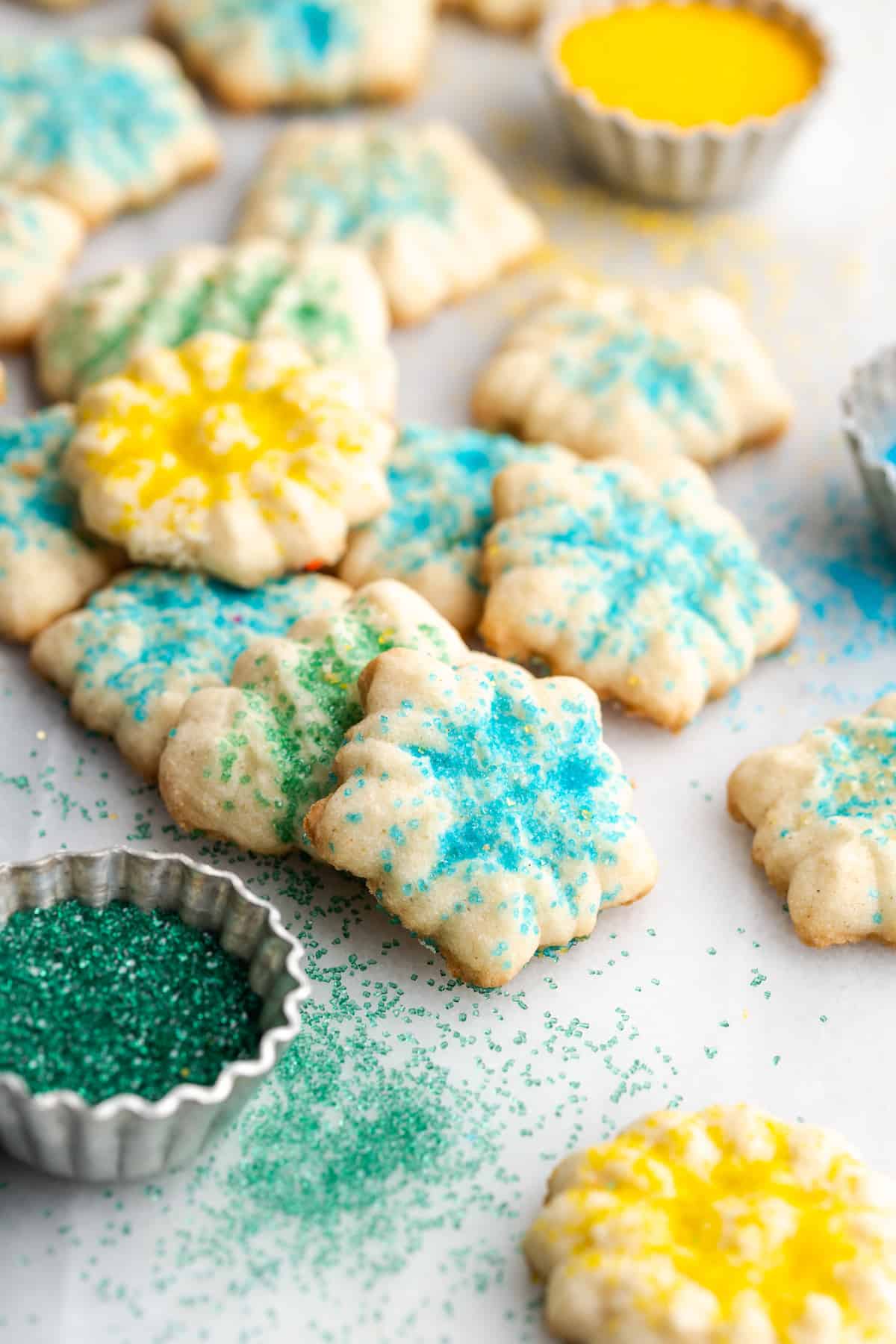 A pile of spritz cookies with various sprinkles on them, next to a bowl of green sprinkles and a bowl of yellow sprinkles