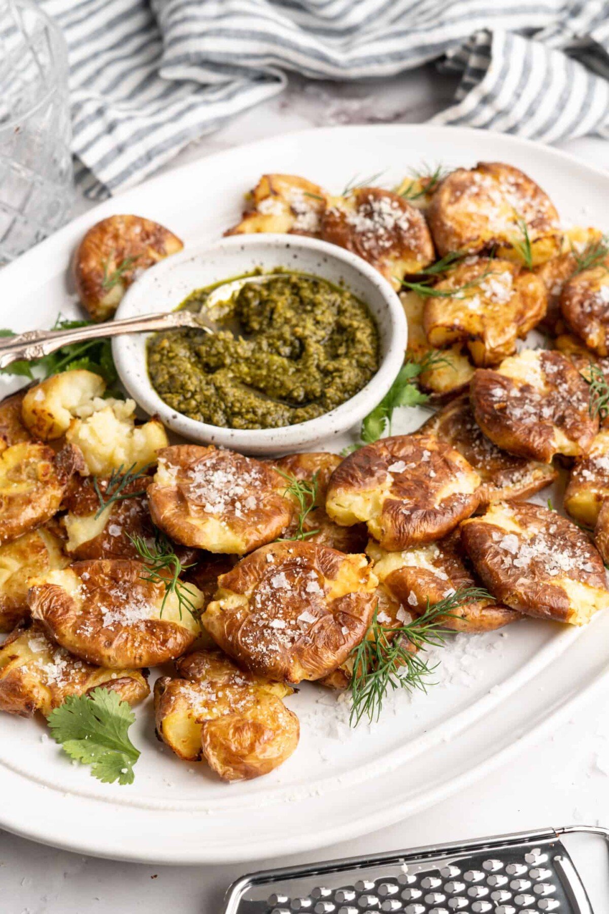 A tray full of smashed potatoes, topped with cheese, with a bowl of pesto with a spoon in it