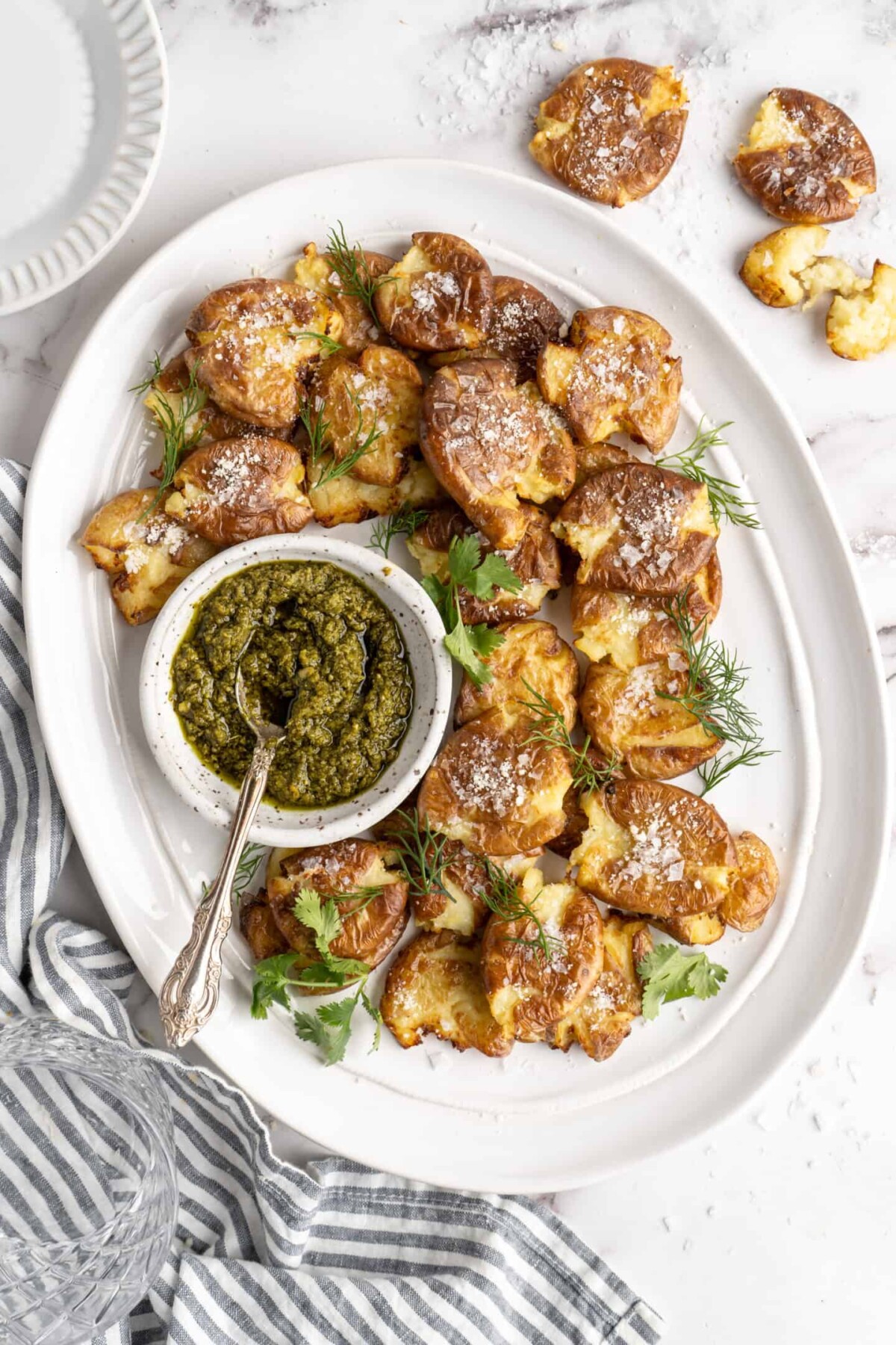 Overhead view of a tray of smashed potatoes garnished with vegan cheese and parsley, with a bowl of pesto with a spoon in it