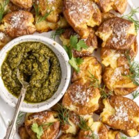 Close up of a tray of smashed potatoes with a bowl of pesto, with a spoon in the pesto.
