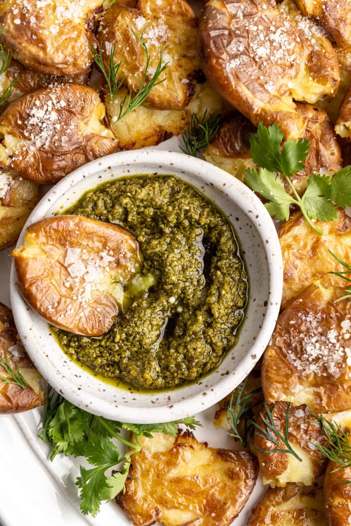 A smashed potato resting in a bowl of pesto, on a plate full of smashed potatoes