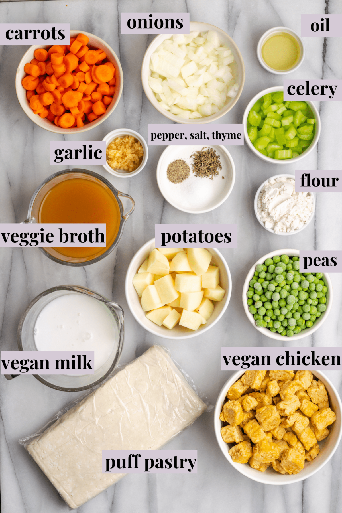 Overhead view of the labeled ingredients for vegan chicken pot pie casserole: a bowl of carrots, a bowl of onions, a bowl of oil, a bowl of celery, a bowl of vegetable broth, a bowl of garlic, a bowl of salt, pepper, and thyme, a bowl of flour, a bowl of peas, a bowl of potatoes, a bowl of vegan milk, a bowl of vegan chicken, and a piece of puff pastry