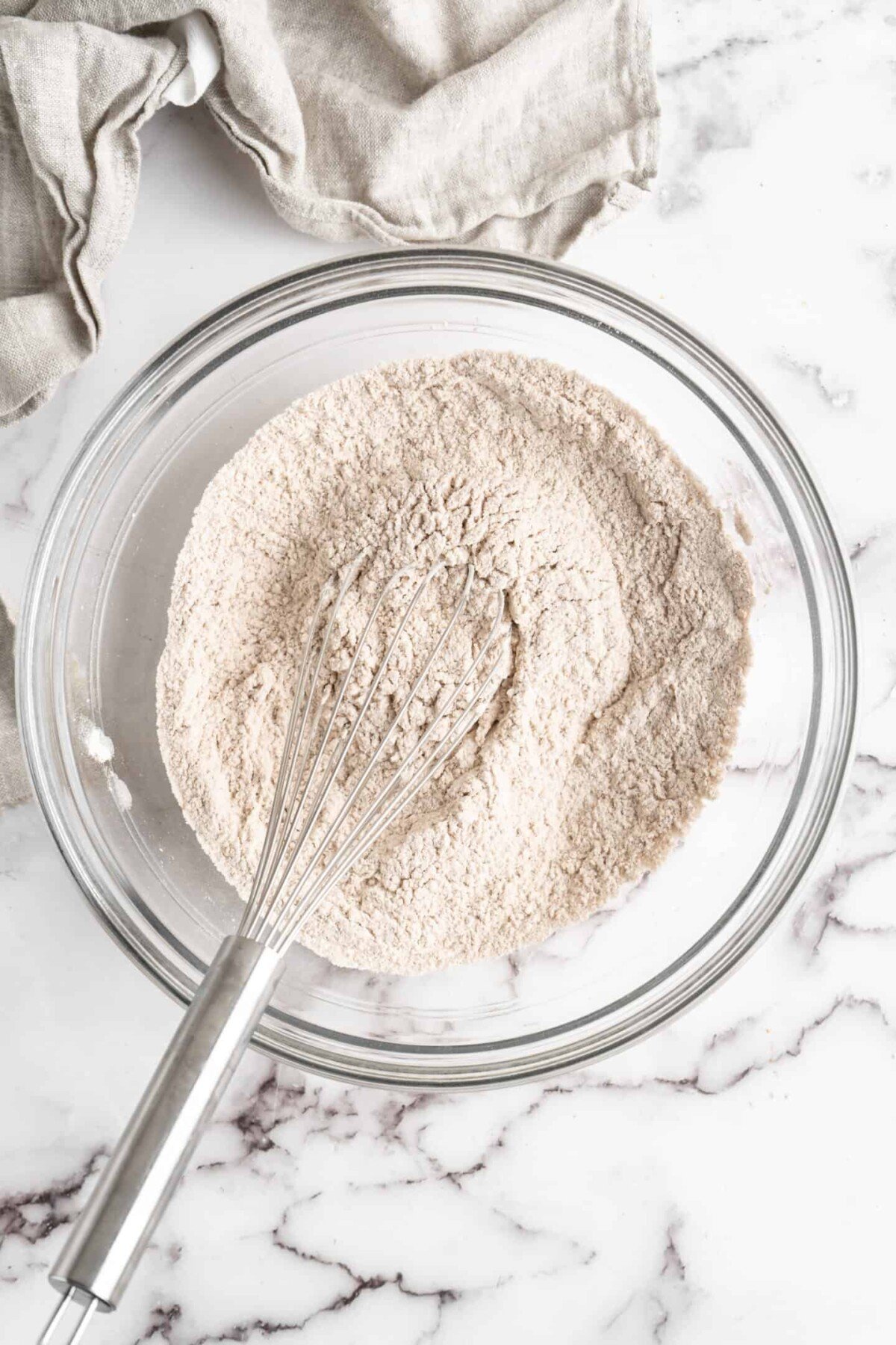 A glass mixing bowl full of flour and spices, with a whisk