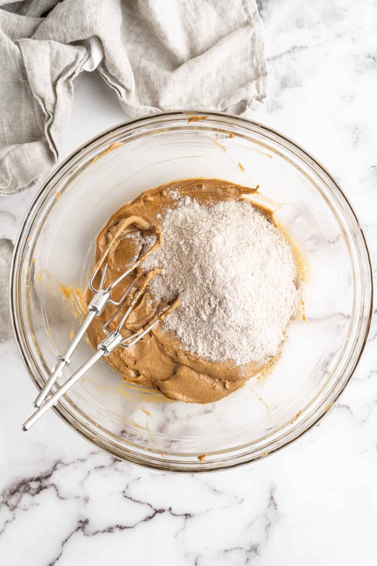 A glass mixing bowl with the wet ingredients for gingerbread men dough beat together, with a pile of dry ingredients on top, and two electric beater blades in the bowl