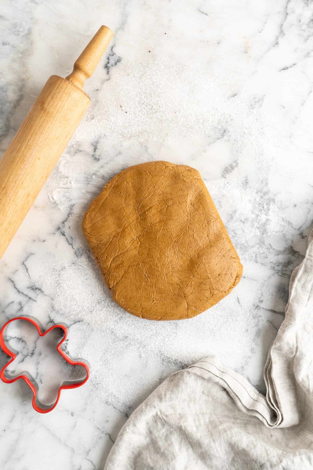 A think disk of gingerbread men dough on a marble counter, next to a kitchen towel, a gingerbread men cookie cutter, and a rolling pin