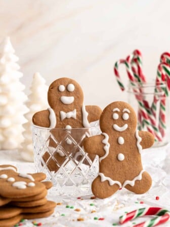 Two gingerbread men standing upright, one in a glass cup, next to a stack of gingerbread men, with a cup of candy canes in the background