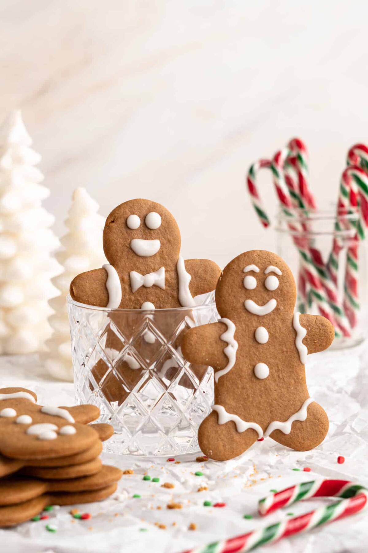 Two gingerbread men standing upright, one in a glass cup, next to a stack of gingerbread men, with a cup of candy canes in the background
