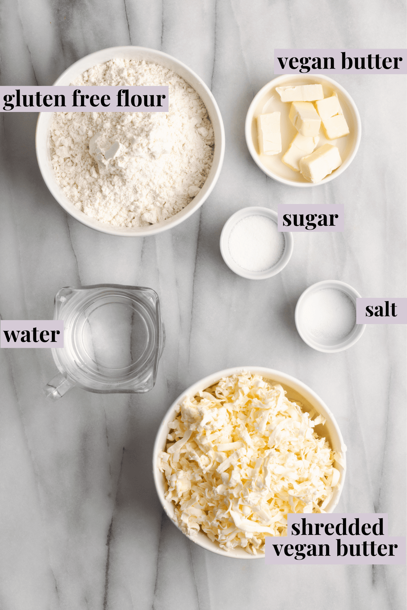 Overhead view of the ingredients for vegan puff pastry: a bowl of gluten-free dough, a glass of water, a bowl of salt, a bowl of sugar, a bowl of cubed vegan butter, and a bowl of shredded vegan butter