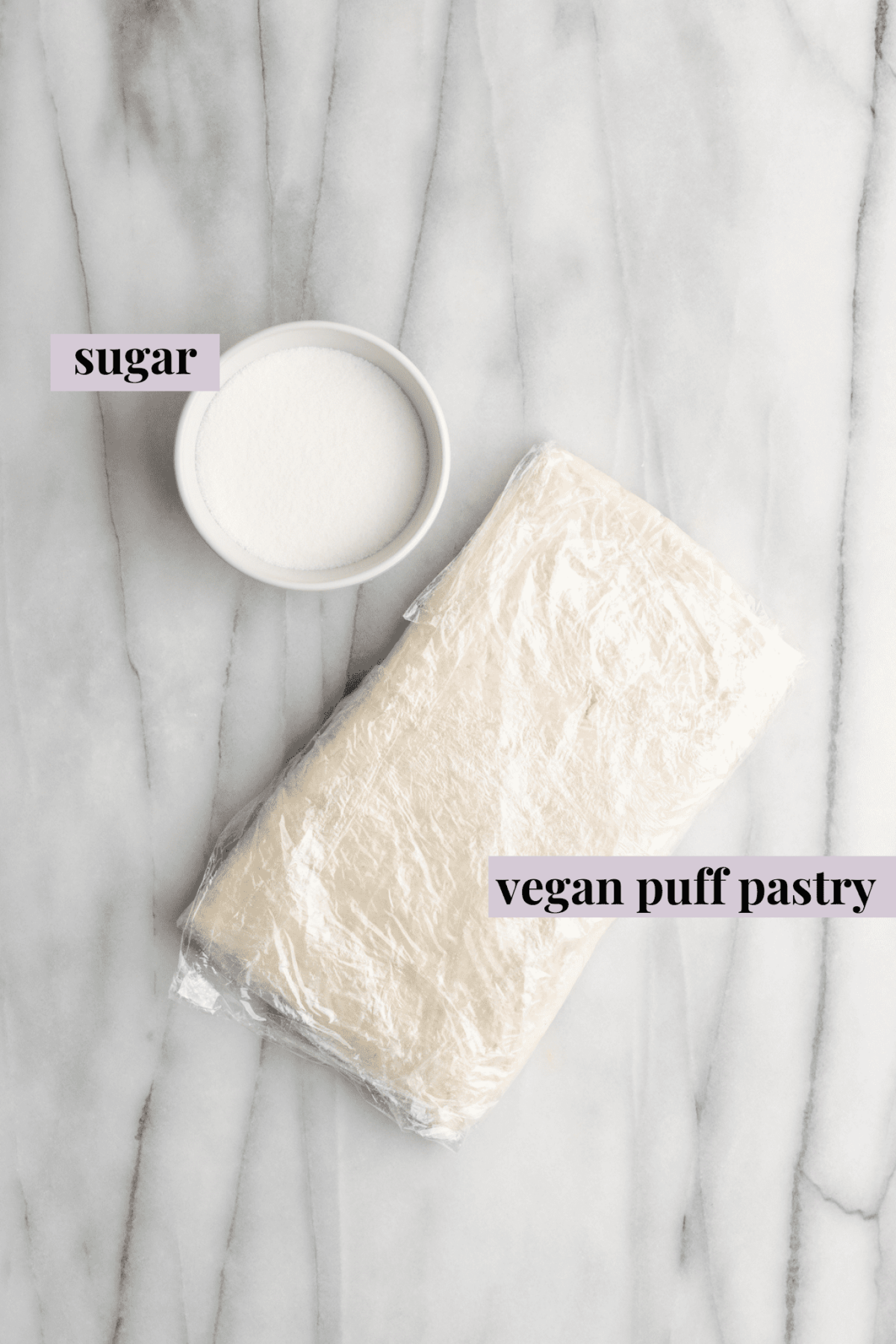 Overhead view of the labeled ingredients for vegan puff pastry palmiers: a bowl of sugar, and a piece of vegan puff pastry wrapped in plastic wrap