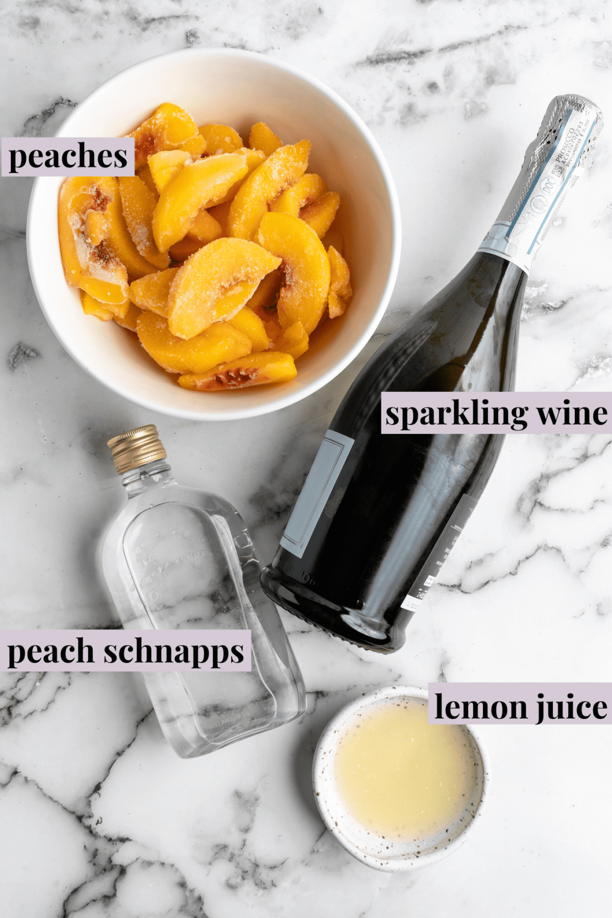 Overhead view of the labeled ingredients for peach bellinis: a bowl of peach slices, a bowl of lemon juice, a bottle of sparkling wine, and a bottle of peach schnapps