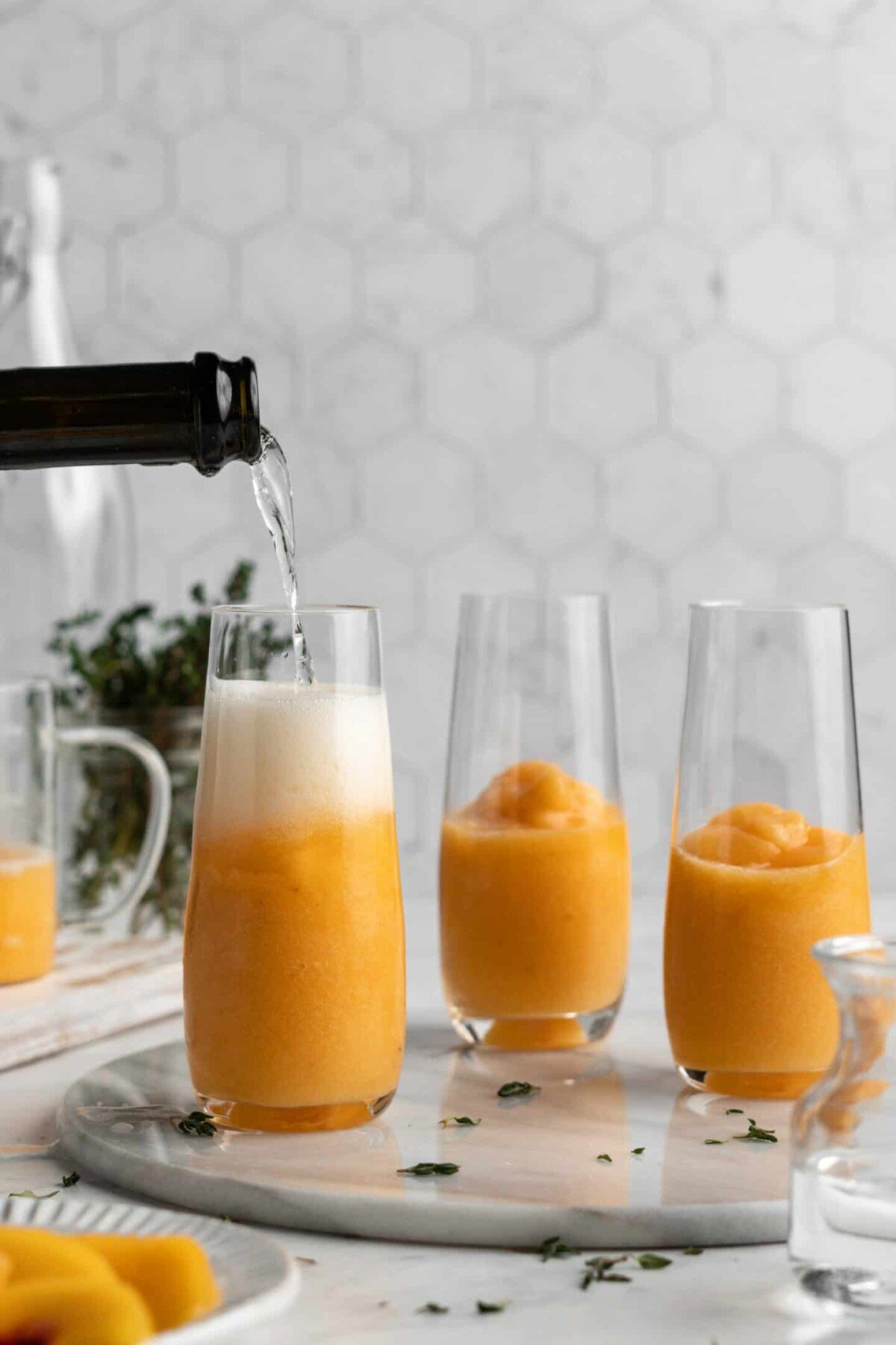 A glass filled halfway with peach puree with a bottle of sparkling wine being poured into it, with two glasses of peach puree in the background