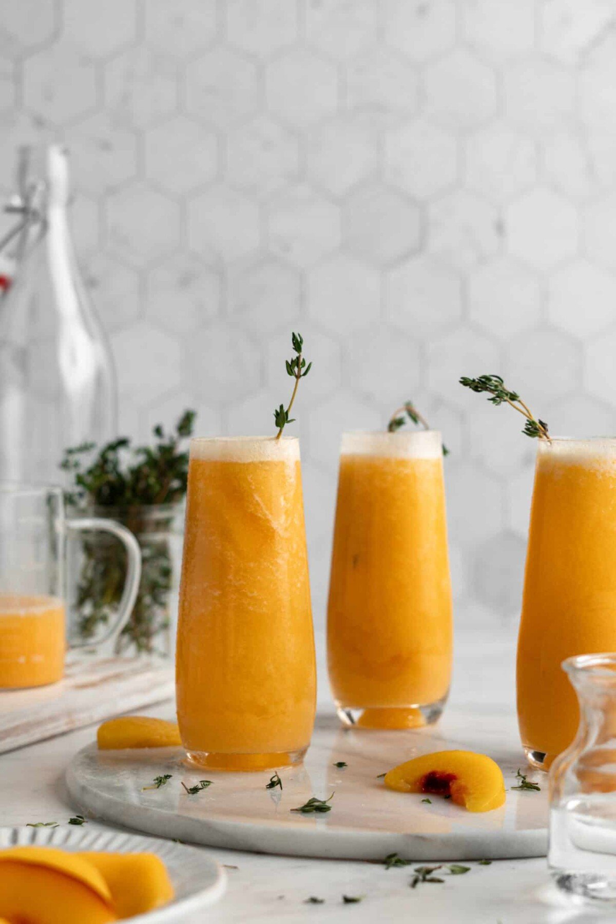 Three peach bellinis with herbs and a pitcher in the background, and herbs and peach slices around them