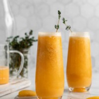 Two glasses of peach bellinis, surrounded by herbs and peach slices, with a sprig of herbs in them