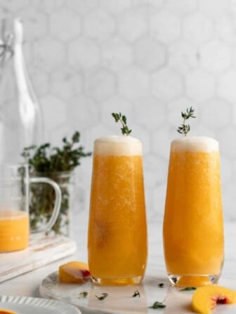 Two peach bellinis with sprigs of herbs in them, surrounded by peach slices and herbs