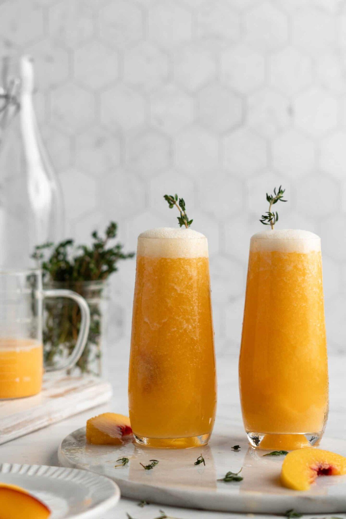 Two peach bellinis with sprigs of herbs in them, surrounded by peach slices and herbs