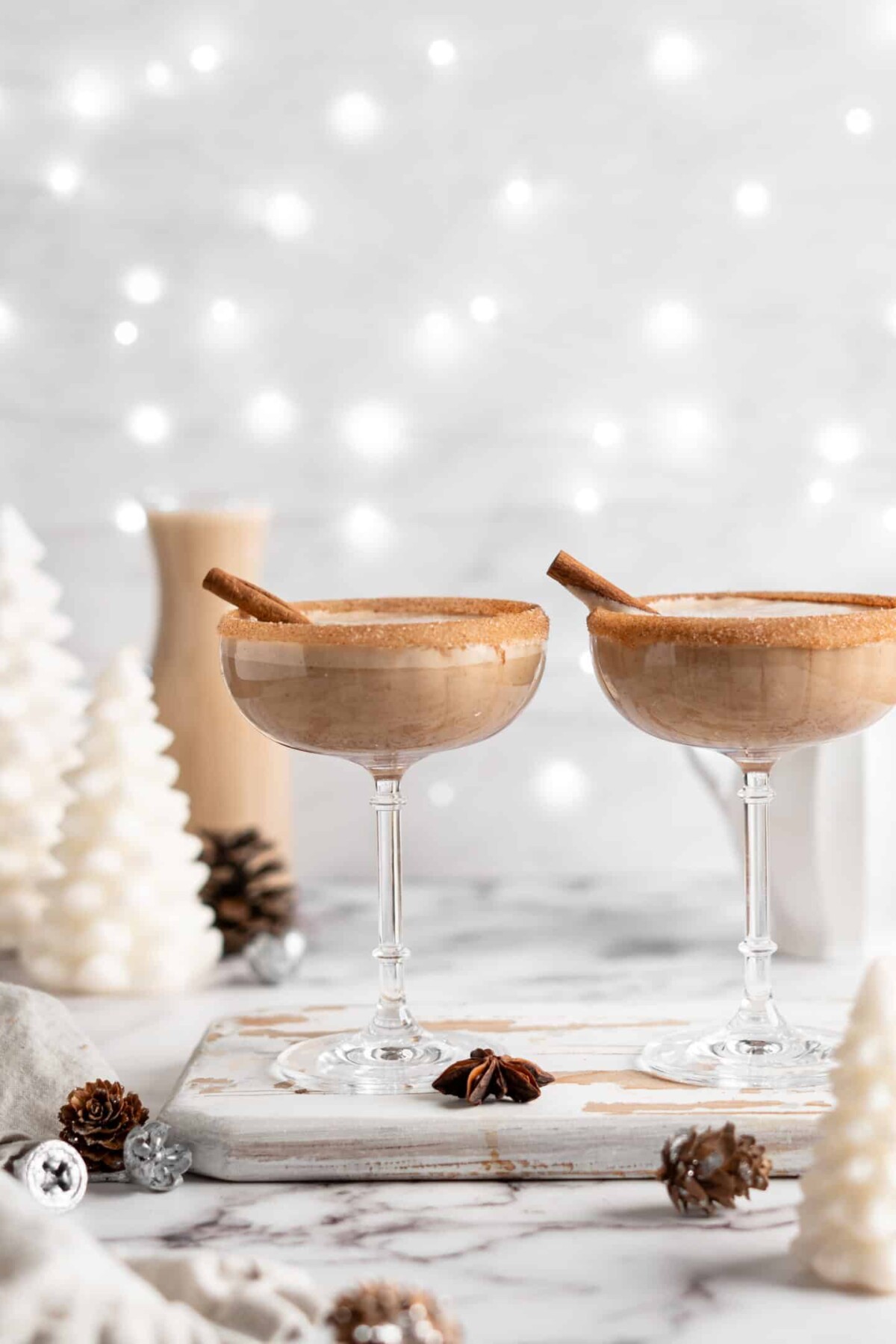 Two cocktail glasses filled with vegan eggnog, with cinnamon sugar rims and cinnamon sticks sticking out of them, surrounded by star anise pods and white Christmas decorations