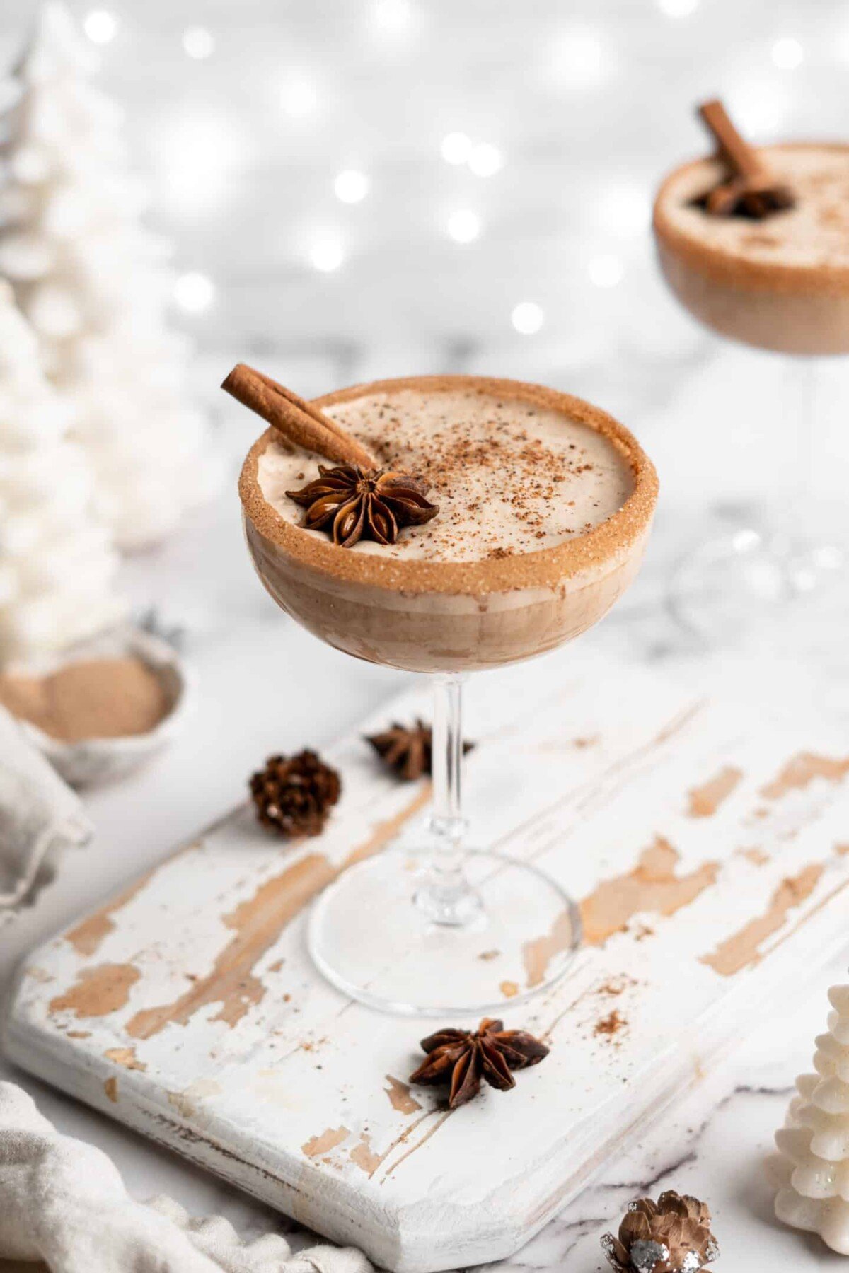 A cocktail glass filled with vegan eggnog, garnished with a cinnamon stick, ground cinnamon, and a star anise pod, with a cinnamon sugar rim, with Christmas lights and another glass of eggnog in the back