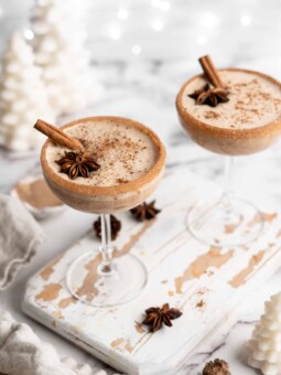 Overhead view of two cocktail glasses with vegan eggnog, topped with ground cinnamon, whole cinnamon sticks, and whole star anise, with cinnamon sugar rims, surrounded by star anise and white Christmas decorations