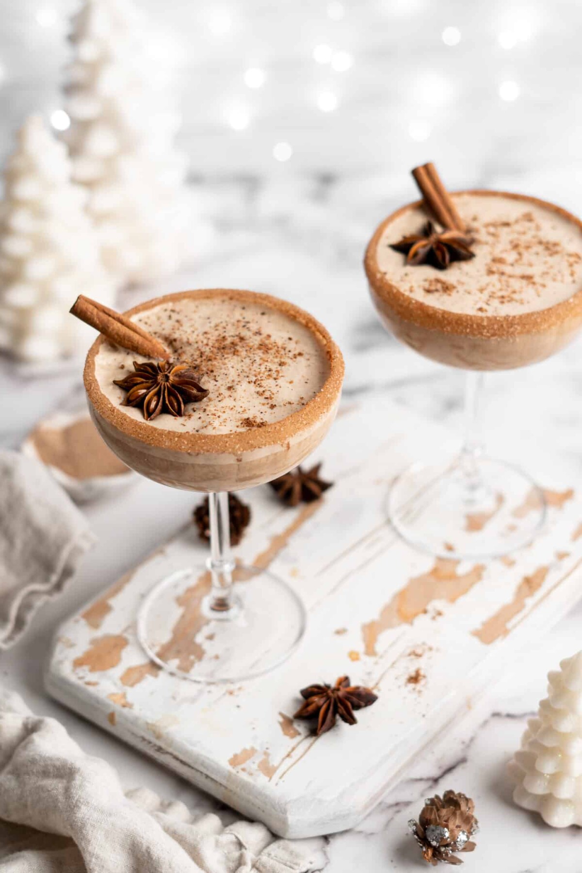 Overhead view of two cocktail glasses with vegan eggnog, topped with ground cinnamon, whole cinnamon sticks, and whole star anise, with cinnamon sugar rims, surrounded by star anise and white Christmas decorations