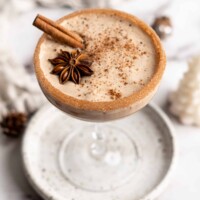A cocktail glass on a plate filled with vegan eggnog, garnished with a cinnamon stick, a star anise pod, and ground cinnamon, with a cinnamon sugar rim