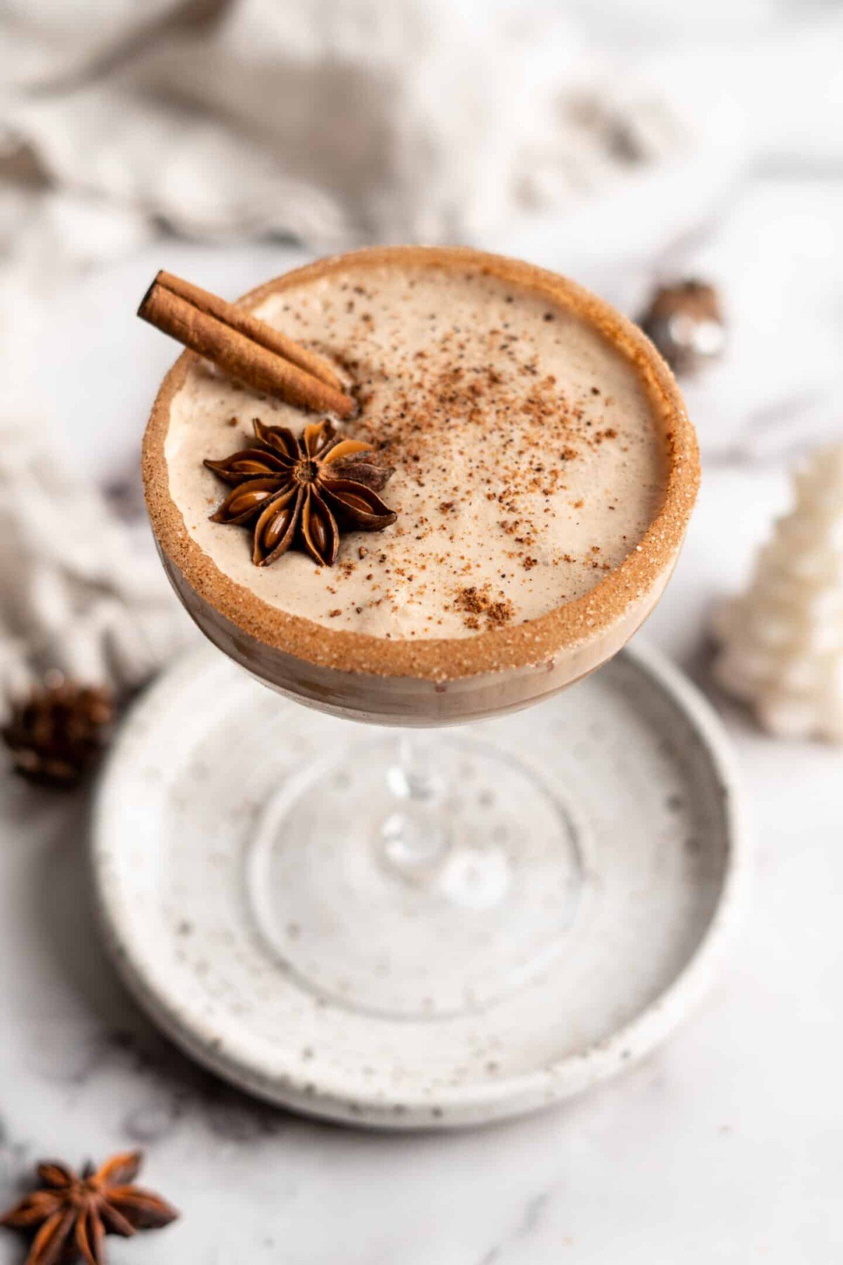 A cocktail glass on a plate filled with vegan eggnog, garnished with a cinnamon stick, a star anise pod, and ground cinnamon, with a cinnamon sugar rim