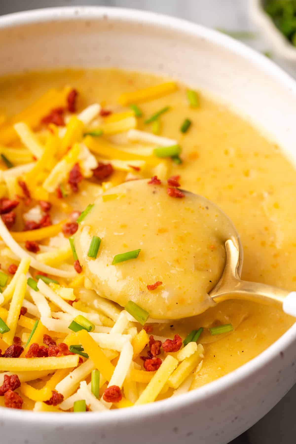 Close up of a spoon taking a spoonful of potato soup out of a bowl of potato soup topped with chives, vegan cheese, and vegan bacon bits