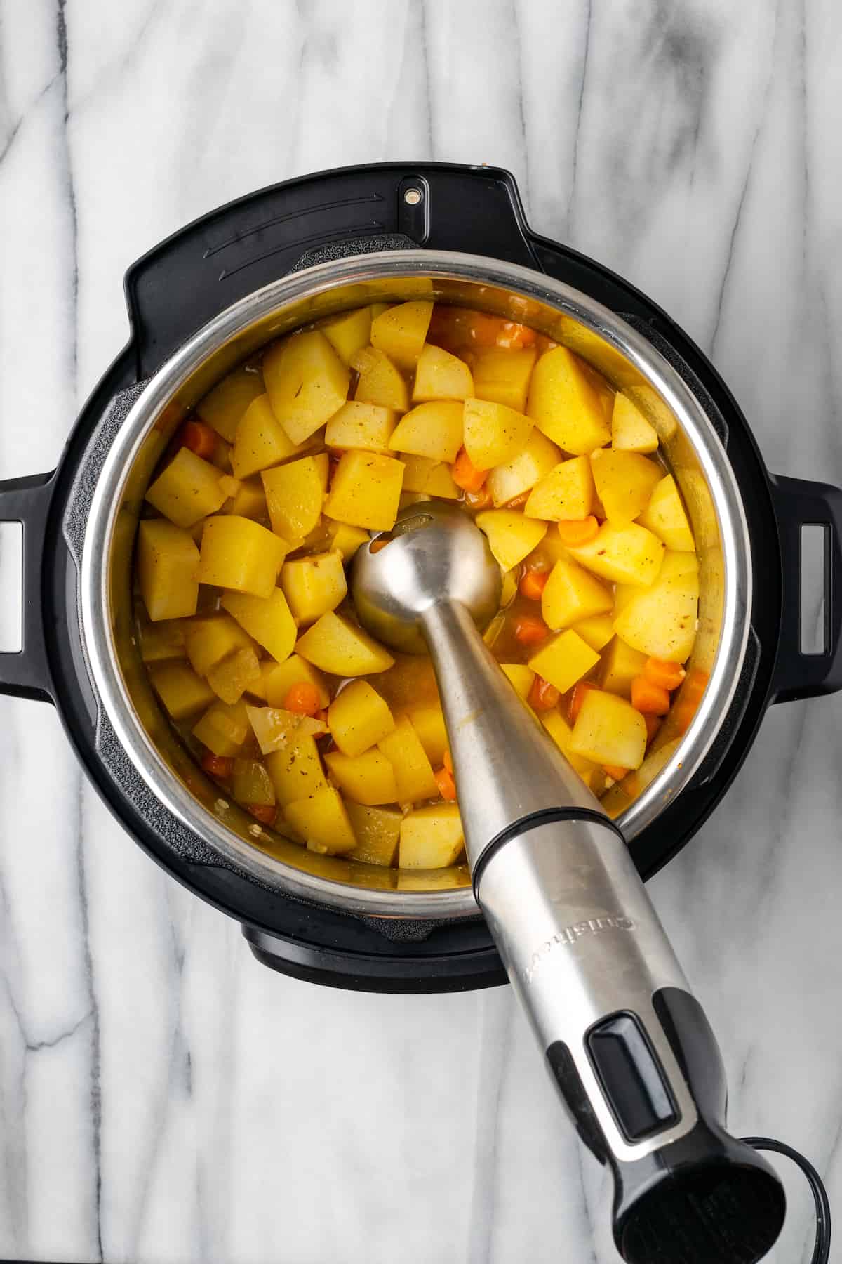 An immersion blender in an Instant Pot with cooked potatoes, carrots, onions, and veggie broth