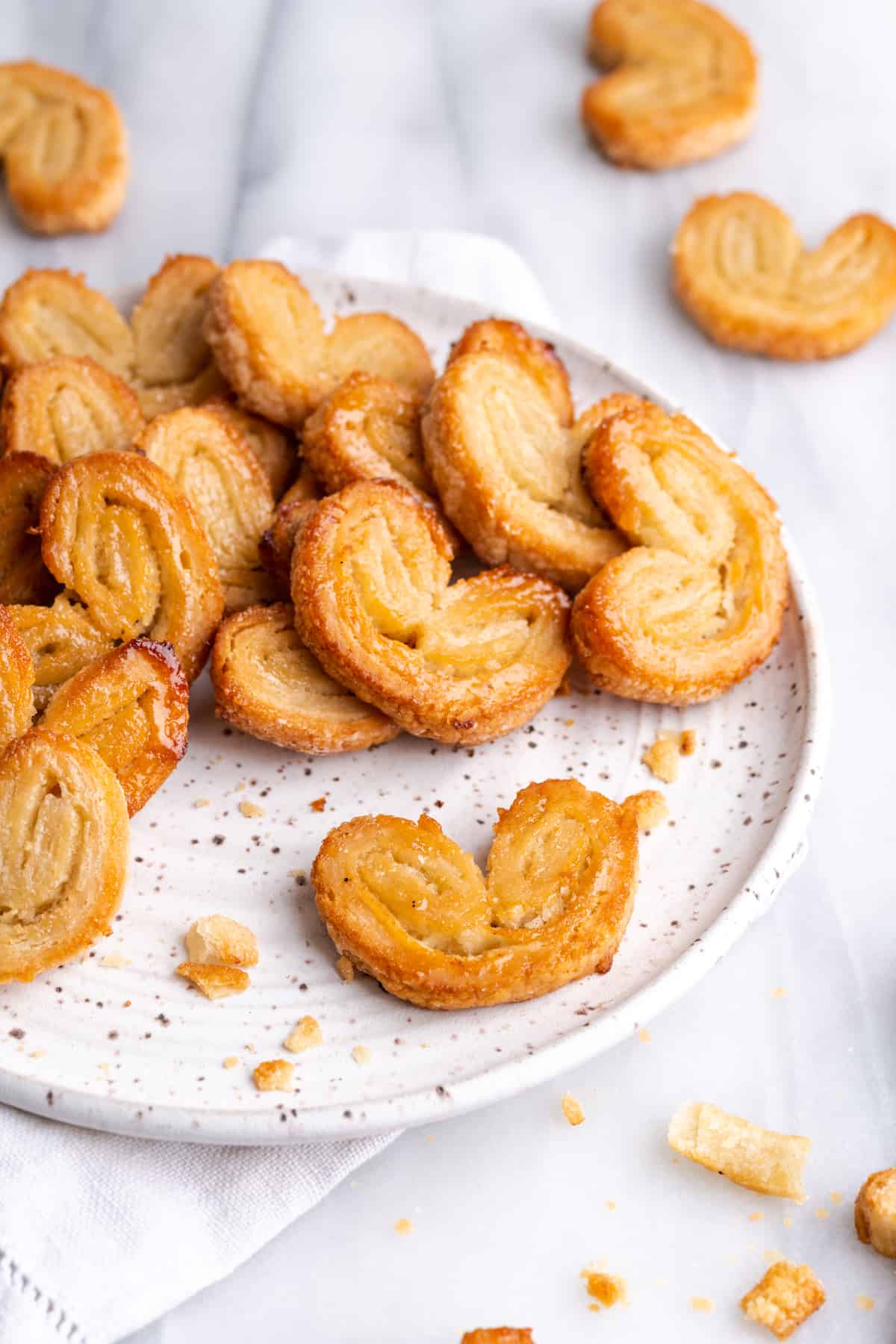 A plate with a pile of puff pastry palmiers, one palmier at the front, and lots of palmiers surrounding it