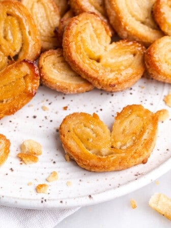 A plate with one puff pastry palmier at the front, and a pile of them at the back