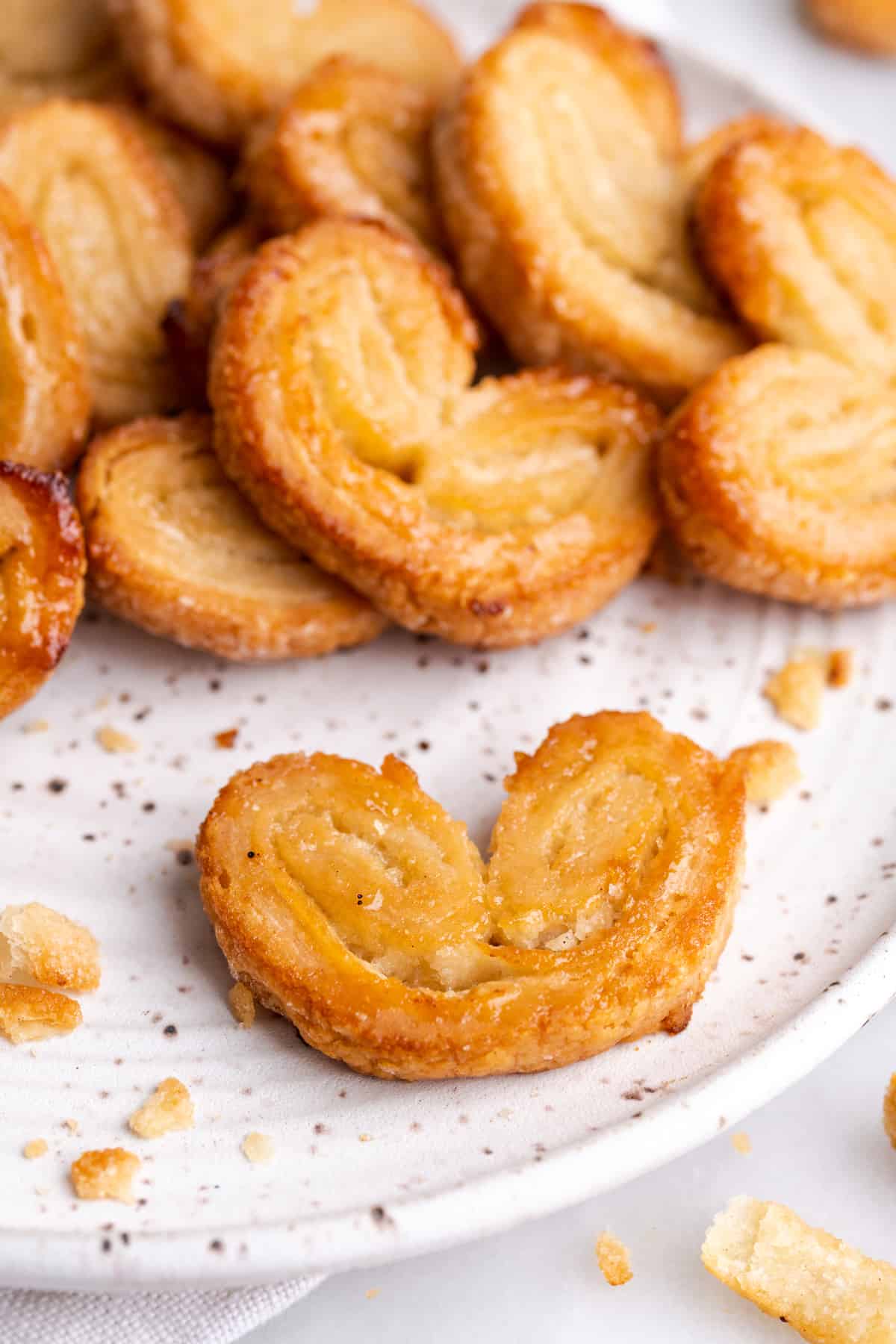 Close up of a puff pastry palmier on a plate, with a pile of palmiers behind it