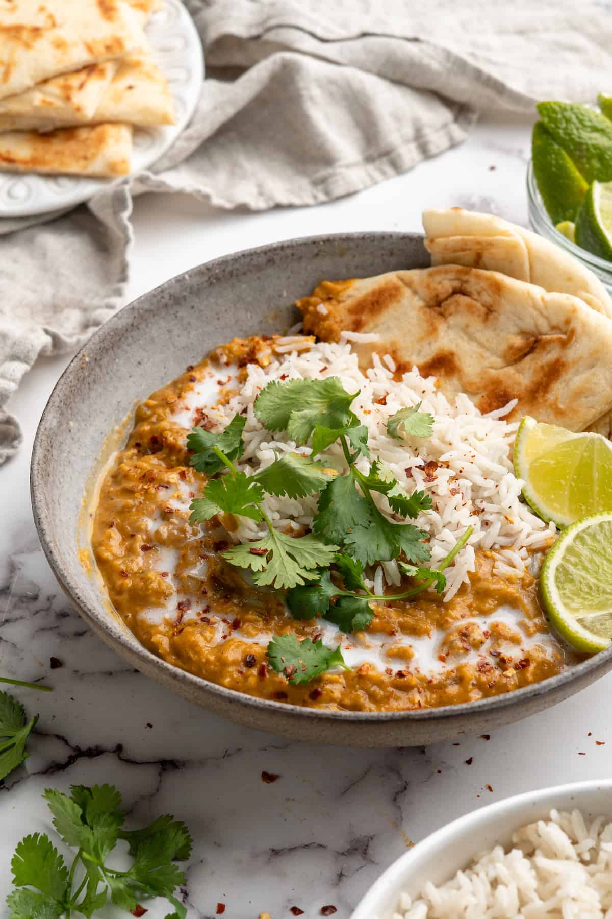 Red lentil curry in bowl with lime wedges, cilantro, rice, and naan