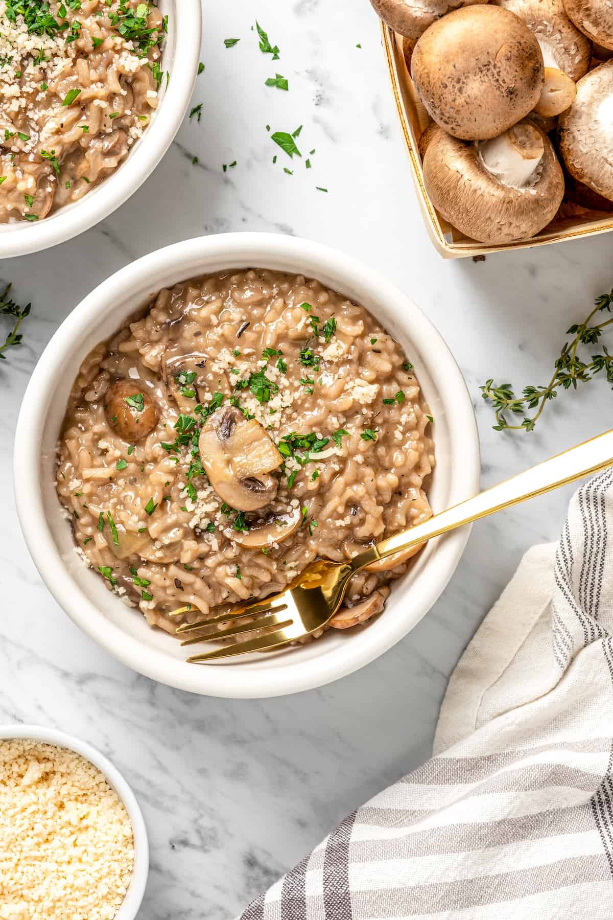 Overhead view of vegan risotto in bowl with carton of mushrooms, tea towel, herbs, and bowl of parmesan