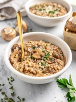 Two bowls of vegan Instant Pot risotto with mushrooms, with fresh herbs and mushrooms on tabletop