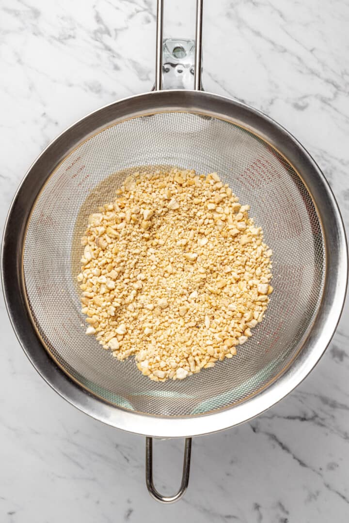 Overhead view of ground cashews in mesh strainer set over liquid measuring cup