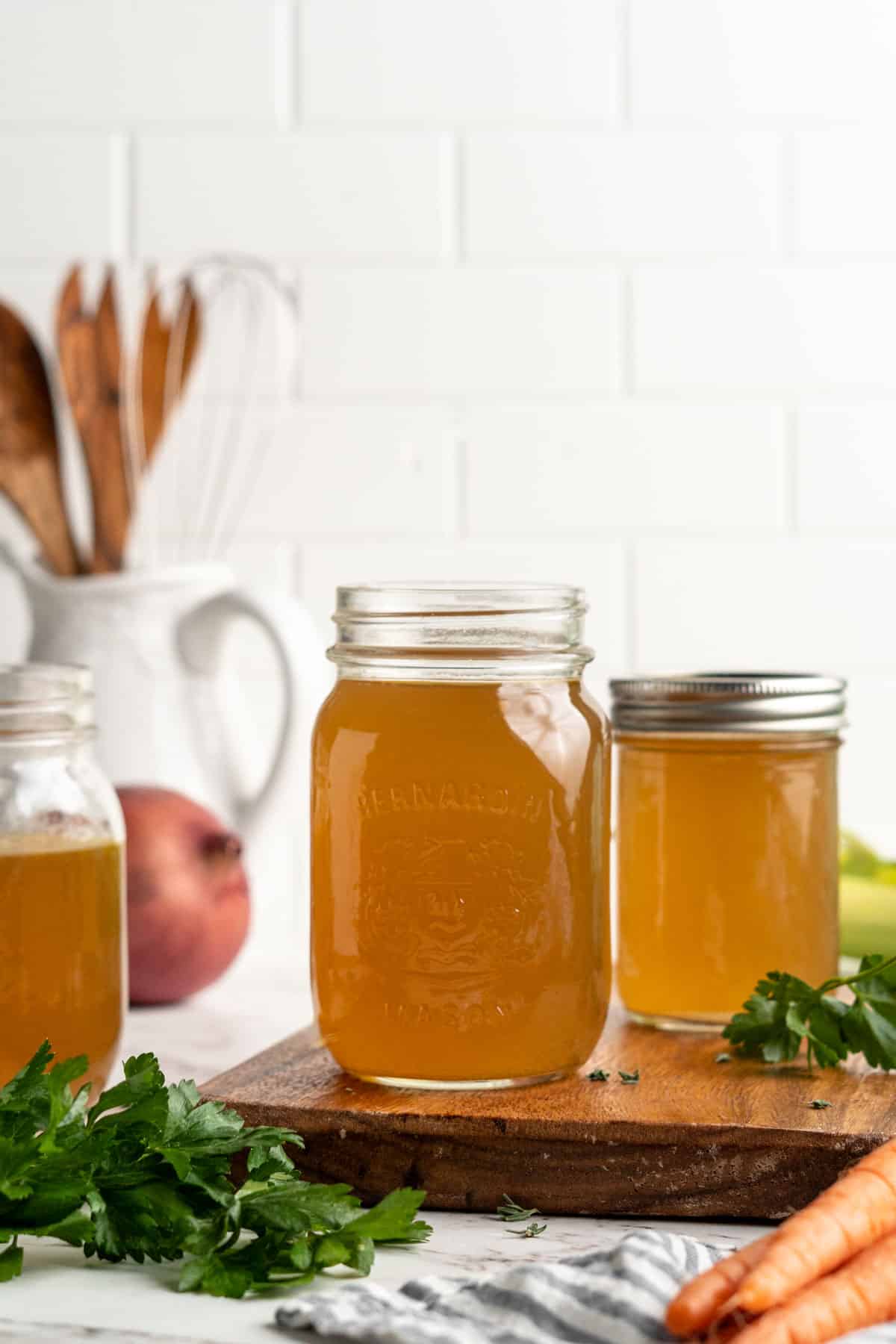 Three jars of homemade vegetable broth, surrounded by broth ingredients