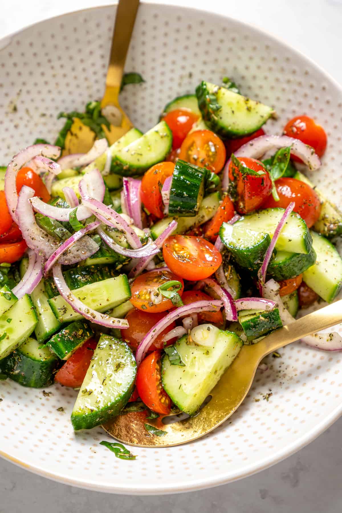 Two gold serving spoons in bowl with vegan cucumber and tomato salad