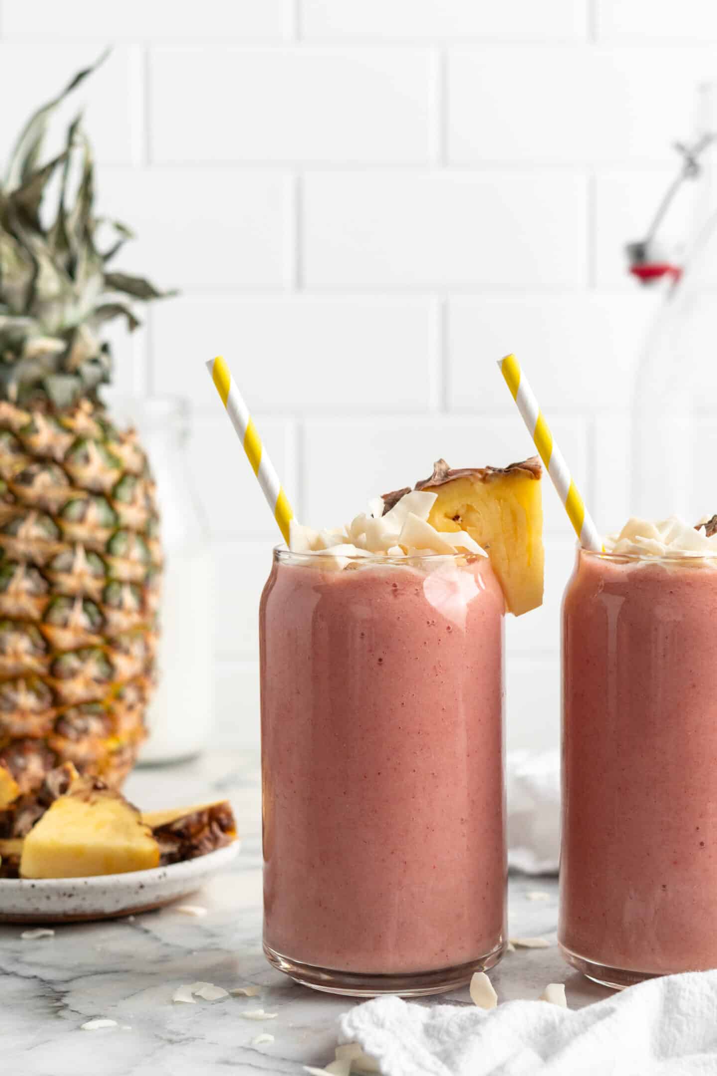 Two strawberry pineapple smoothies in glasses with straws, garnished with coconut flakes and pineapple wedge
