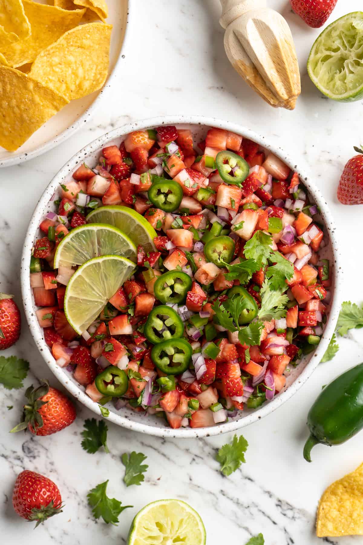 Overhead view of strawberry salsa in bowl, surrounded by strawberries, lime, peppers, and cilantro on marble countertop