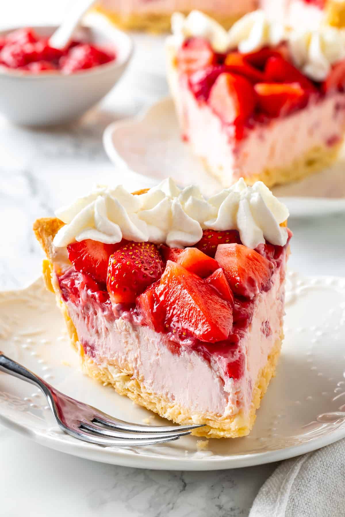 Slice of strawberry cream pie on plate with fork, with another slice of pie in background