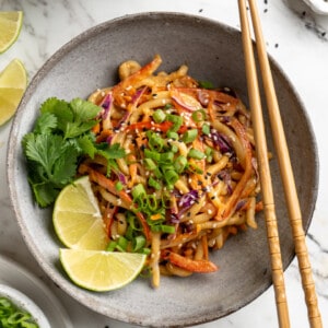 Overhead view of Thai peanut noodles in bowl with lime wedges, cilantro, and green onions for garnish