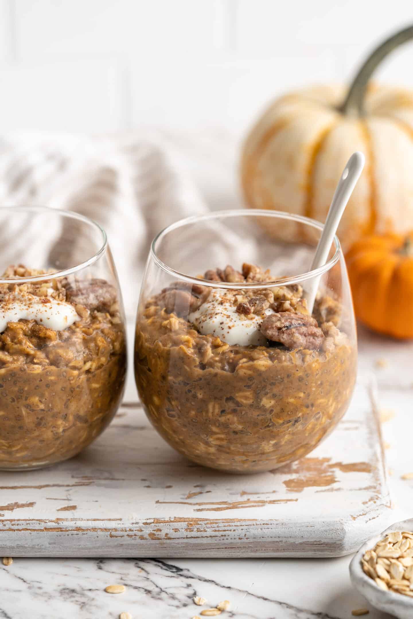 two jars of pumpkin pie overnight oats with spoon inside one and pumpkins in background 45 degree angle