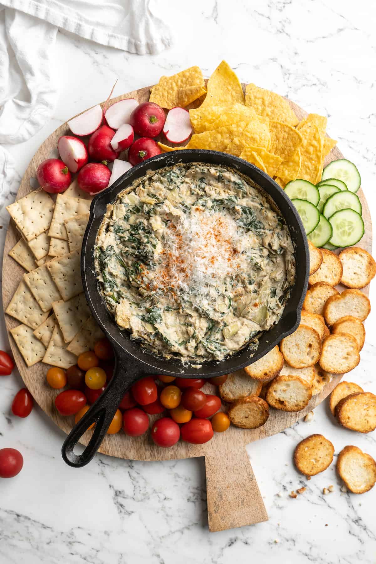 Overhead view of snack board with skillet of spinach artichoke dip, chips, vegetables, and crackers